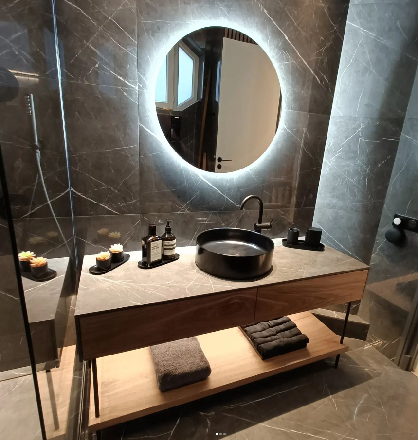 Meuble de salle de bain en bois noyer sur mesure avec vasque noire, miroir rond éclairé et carrelage en marbre gris.